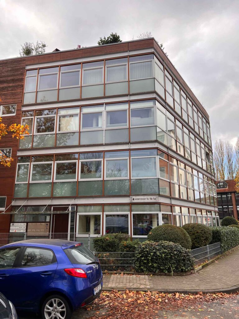 Ein modernes, vierstöckiges Gebäude mit großen Glasfenstern und braunen Rahmen steht in DE, umgeben von Büschen. Ein blaues Auto ist auf der Straße davor geparkt, und an einem Baum in der Nähe ist Herbstlaub zu sehen. Kontaktieren Sie uns für weitere Details.
